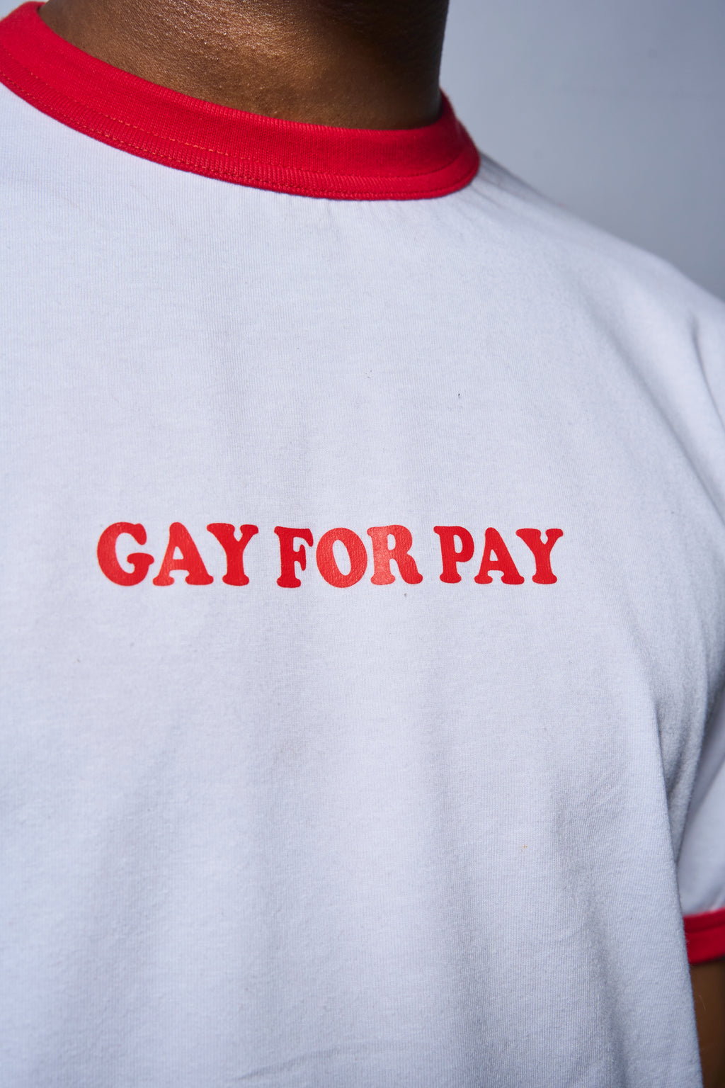 Close-up of a person wearing a white ringer T-shirt with red trim at the collar and sleeve cuffs, featuring the slogan 'GAY FOR PAY' printed in red across the chest.